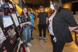 Three students stand in front of a poster board, placed on a table. A museum visitor stands in front of them looking at the poster board.