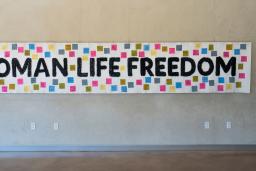 A white banner hangs on a wall, bearing the words "Woman, Life, Freedom" in black. Small coloured squares with text are attached to the banner around the text.