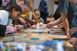 Un groupe d’enfants et de jeunes peint avec de petits pinceaux, de part et d’autre d'une longue table. Au premier plan, il y a des peintures de différentes couleurs.