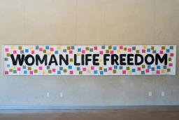 Une banderole blanche est accrochée à un mur, portant les mots "Femme, Vie, Liberté" en noir. De petits carrés de couleur avec du texte sont attachés à la bannière autour du texte.