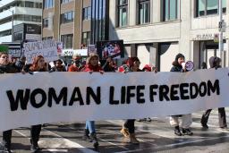 Six personnes portent une longue bannière blanche sur laquelle on peut lire les mots anglais en lettres majuscules noires "FEMME VIE LIBERTÉ" dans une rue de la ville de Toronto. Derrière elles, un défilé de femmes porte d'autres pancartes de protestation pour la Journée internationale des femmes.