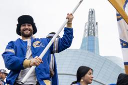 Un homme souriant dans une foule, tenant un grand drapeau jaune avec un « W » bleu et blanc au centre. L’homme porte une veste bleue des Blue Bombers de Winnipeg et se tient devant le Musée canadien pour les droits de la personne.