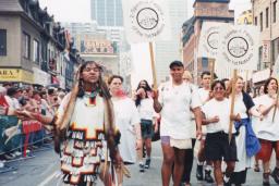 Un groupe de personnes portant des pancartes sur lesquelles on peut lire « 2-Spirited People of the 1st Nations » marchent au centre d’une rue de la ville, tandis que des gens regardent derrière des barrières de part et d’autre de la rue.