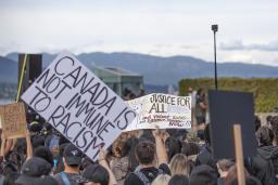 Une foule de personnes faisant dos à la caméra. Certains membres de la foule tiennent des pancartes au-dessus de leur tête qui disent, en anglais : « Je ne peux pas respirer #BLM », « Le Canada n'est pas à l'abri du racisme », et « Justice pour tous : mettez fin au racisme violent, subtil et systémique maintenant!!! ».