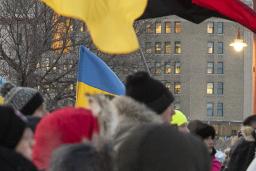 Une foule de personnes emmitouflées se tient debout dans une rue de ville pendant l’hiver, tenant de multiples drapeaux ukrainiens bleus et jaunes ainsi qu’un drapeau canadien. Le rassemblement semble être une manifestation en appui à l’Ukraine. De grands édifices à bureaux et des feux de circulation sont à l’arrière-plan sous un ciel couvert.