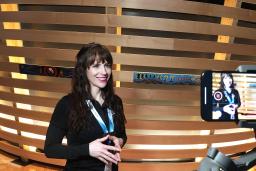 Une femme souriante à la peau claire se fait filmer avec un téléphone portable. Elle porte un cordon autour du cou sur lequel on peut lire "Laissez-vous inspirer" et se tient devant un élément d'exposition fait de bandes de bois horizontales, certaines décorées d'art autochtone.