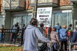 Une file de personnes attend à l’extérieur d’un bâtiment. Des panneaux indiquent qu’il s’agit d’une banque alimentaire. Un drapeau canadien flotte au-dessus d’elles.