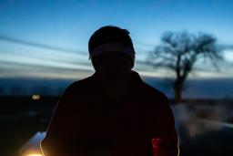 Au premier plan, la silhouette d’une personne qui semble courir vers nous, devant une voiture dont les phares sont allumés. À l’arrière-plan, un ciel bleu pâle avec la silhouette d’un arbre et des nuages longs et fins qui traversent l’horizon.