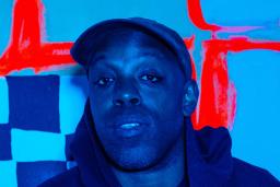 Image aux tons bleus d’un homme noir portant une casquette et un chandail à capuchon foncé devant un fond à carreaux avec d’épaisses lignes rouges.