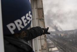 Une personne portant un casque bleu foncé marqué du mot « PRESS » montre, depuis la fenêtre d’un balcon surplombant une route, une colonne de fumée à la suite d’une explosion.