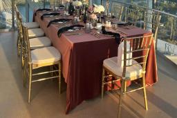 Nichée dans un coin de la plateforme d’observation de la tour de verre, une table somptueusement décorée avec une nappe rouge vin, des compositions florales et de la vaisselle, et des chaises dorées.