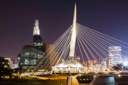 Photo de nuit d’un édifice étonnant, surmonté d’une tour illuminée, près d’un pont moderne lui aussi illuminé, qui se reflète dans l’eau de la rivière dans le bas de la photo.