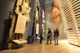 Cinq personnes regardent des expositions imposantes dans la galerie « Perspectives autochtones » du Musée. La plus proche est en bois et représente des arbres, des animaux et une plaque. Derrière les personnes, on aperçoit un théâtre de forme circulaire, construit avec des lattes de bois courbées.