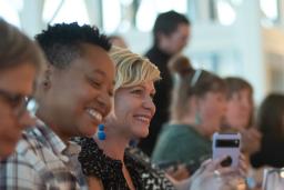 Un groupe de personnes est assis à une longue table, riant et appréciant un repas ensemble. L’accent est mis sur deux personnes qui sourient joyeusement – l’une portant une chemise à carreaux et l’autre un haut noir à motifs, avec des verres et des assiettes soigneusement disposés devant elles.