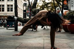 Une femme vêtue d'un coton ouaté gris et d'un pantalon gris foncé fait du breakdance sur un trottoir pendant la journée. Des arbres et des bâtiments sont à l'arrière-plan.