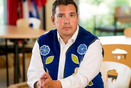 A man wearing a blue vest with flower beadwork, seated with his hands resting on a table.