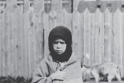 Une jeune fille se tient face à la caméra avec une expression de tristesse, serrant un châle autour d’elle.