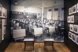 Une exposition muséale qui comprend de petits pupitres d’école devant un grand fond photographique d’enfants autochtones assis à leur pupitre dans une classe.