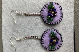 Une paire de boucles d'oreilles en feutre violet clair avec des bordures cousues violet foncé. Au milieu des boucles d'oreilles se trouvent des fleurs en perles.