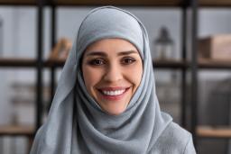 Une femme portant un hidjab bleu sourit vers l'objectif. Il y a une étagère de livres en arrière-plan.