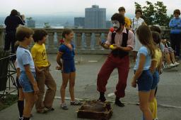 Un homme au visage peint jongle en plein air, sous le regard d'enfants souriants. Une silhouette de ville apparaît au loin derrière lui.