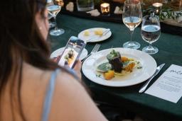 Une femme à la peau claire et aux longs cheveux bruns utilise son téléphone portable pour prendre une photo d’une assiette de nourriture gastronomique posée sur une table.