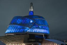 L’extérieur du Musée canadien pour les droits de la personne est photographié de nuit. Le bâtiment est éclairé de l’intérieur par des lumières vives d’un bleu profond.