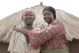 Deux femmes debout devant une hutte avec un toit de paille, regardent la caméra. La femme plus à droite sourit en serrant l’autre femme dans ses bras.