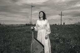 Une femme vêtue d'une longue robe blanche se tient dans un champ en tenant une bandura, un instrument folklorique ukrainien à cordes pincées.