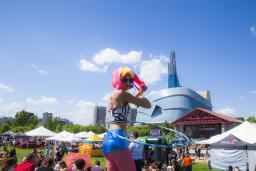Une personne portant une perruque de couleur vive et utilisant un hula-hoop lors d’un festival en plein air; on voit le Musée dans l’arrière-plan.