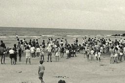 Une photographie en noir et blanc d’une foule de personnes, la plupart debout, sur une plage.