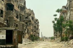 Une rue de ville abandonnée où les fenêtres des bâtiments ont été détruites, des gravats sont empilés sur la route et un camion est abandonné avec ses portes arrière ouvertes.