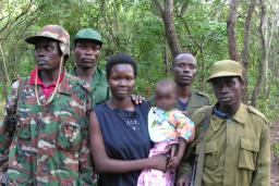 Une femme tenant un bébé, tandis que quatre hommes en tenue militaire se tiennent à côté et derrière elle. Ils sont tous debout devant une forêt et posent pour la photo.