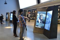 Un homme et une femme sont debout et regardent un écran vidéo en deux parties dans une galerie. L'homme a le bras allongé vers l'écran.