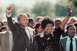 Nelson Mandela, un homme noir âgé aux cheveux grisonnants, forme un poing levé avec sa main droite et le tient au-dessus de sa tête. De sa main gauche, il tient la main de Winnie Mandela, une femme noire aux cheveux noirs. Elle sourit et forme un poing levé avec sa main gauche, à l'image du poing levé de Nelson Mandela. Une foule nombreuse est rassemblée derrière eux.