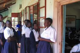 A group of young women and girls in their teens stand together on the front porch of a building. Most are dressed in white blouses and blue skirts, with two girls also wearing blue sweaters. To the right, a doorway reveals a roomful of students sitting at wooden desks and writing on notepads.