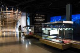 Une galerie de musée contenant une structure circulaire formée de lattes horizontales de bois, des vitrines contenant des objets et des projections sur le mur.