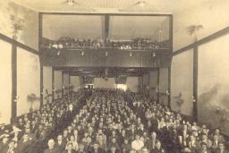 Photo en noir et blanc d’un public de cinéma. La photo est prise de l’avant du cinéma en regardant vers l’arrière, de sorte que les visages du public sont visibles. Des palmiers en pot bordent les murs de chaque côté.