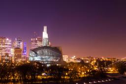 Le panorama d'une ville la nuit. À l'avant-plan, il y a une rivière bordée d'arbres, un bâtiment lumineux en verre et un pont.