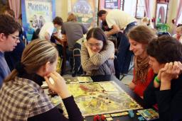 Trois jeunes femmes et deux jeunes hommes sont assis autour d’une table et jouent un jeu de société. En arrière-plan, d’autres personnes jouent à des jeux à d’autres tables.