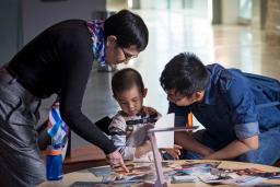 Une femme, un homme et un jeune garçon sont rassemblés autour d’une table circulaire. Plusieurs photographies sont éparpillées sur la table et la femme en montre une au garçon.