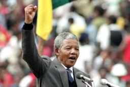 Nelson Mandela, un homme plus âgé à la peau foncée et aux cheveux gris et courts, est debout sur un podium et parle dans deux microphones. Il porte un complet gris et une cravate. Il lève son poing droit au-dessus de sa tête. 