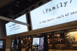 Une femme et un enfant interagissent avec un élément d'exposition numérique dans une galerie de musée. Au-dessus d'eux, sur un grand écran, on peut lire, en anglais, "La famille est l'unité de base de la société."