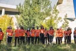 Des personnes portant des t-shirts orange debout devant une rangée d'arbres. À l'avant-plan, on voit des herbes hautes. Sur les t-shirts, de petites empreintes de main sont regroupées en forme de cœur avec le numéro 215+ au-dessus.