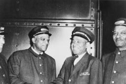 Photo en noir et blanc de quatre hommes en tenue de porteurs ferroviaires. Tous les quatre sourient et les deux du centre se serrent la main.