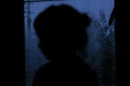 Image sombre de la silhouette d’un jeune enfant contre une fenêtre à travers laquelle on voit des arbres flous et un poteau électrique. La lumière est un ton violacé de crépuscule.