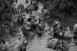 Des femmes, des enfants et des hommes rohingyas marchent dans l’eau d’une rivière qui leur monte jusqu’à la taille. Certaines personnes portent des jeunes enfants alors que d’autres portent des sacs de possessions, dont des articles ménagers.