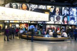 Une grande foule dans une galerie de musée. Il y a un banc courbe au centre de la salle. Dans l’espace, on voit une série de grandes images placées au-dessus d’alcôves qui contiennent chacune une exposition différente.