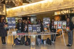 Dans une salle de musée, six étudiants noirs présentent fièrement leur travail sur des affiches et sur une table.