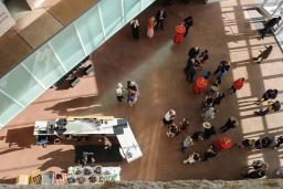 Une vue à vol d'oiseau d'une foule de personnes rassemblées dans un grand espace ouvert du Musée. Il y a quatre tables de cocktail recouvertes de nappes rouges et un bar sur le côté gauche de l'image.